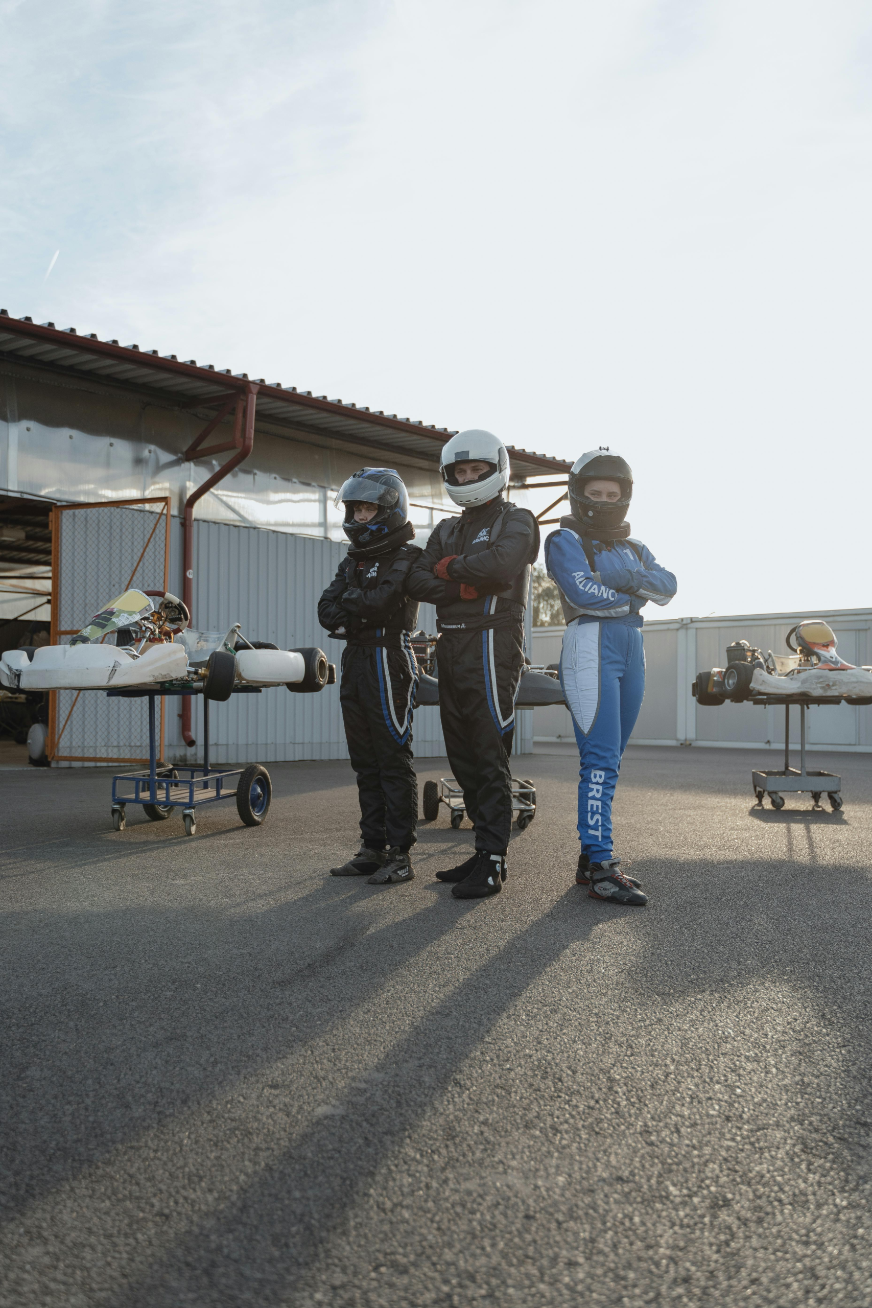 Three professional kart racers in full gear standing confidently outdoors at a racing track.